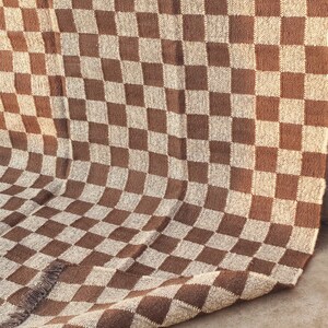 May include: A brown and beige checkered woven textile, possibly a blanket or rug. The pattern is a classic checkerboard design, with alternating squares of brown and beige. The textile has a fringed edge.