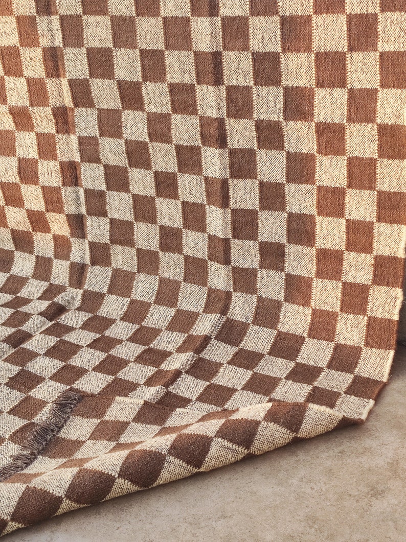 May include: A brown and beige checkered woven textile, possibly a blanket or rug. The pattern is a classic checkerboard design, with alternating squares of brown and beige. The textile has a fringed edge.