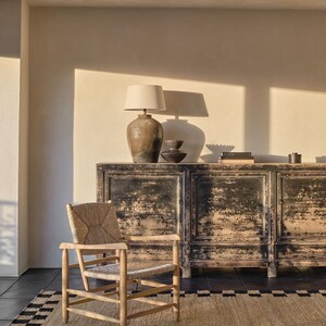 K&ouml;nnte beinhalten: Rustikaler Wohnraum mit einem Holzstuhl mit geflochtener Sitzfl&auml;che, einem schwarzen, abgenutzten Schrank, einer Lampe mit beigem Schirm und einem gemusterten Teppich. Der Raum ist von warmem Sonnenlicht durchflutet und schafft eine gem&uuml;tliche Atmosph&auml;re.