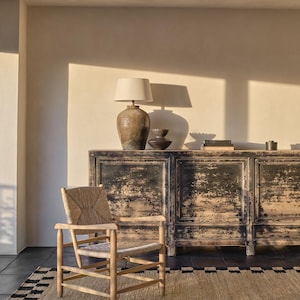 Pu&ograve; includere: Un interno di soggiorno rustico con un mobile nero anticato, una sedia in vimini e un tappeto beige con bordo bianco e nero. Una lampada con paralume beige &egrave; posta sopra il mobile, creando un'atmosfera calda. La stanza ha una palette di colori neutri.