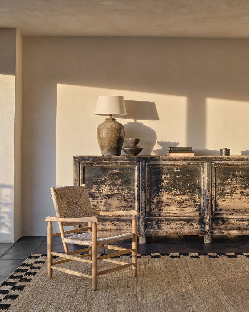 Pu&ograve; includere: Un interno di soggiorno rustico con un mobile nero anticato, una sedia in vimini e un tappeto beige con bordo bianco e nero. Una lampada con paralume beige &egrave; posta sopra il mobile, creando un'atmosfera calda. La stanza ha una palette di colori neutri.