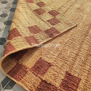 Pu&ograve; includere: Un tappeto in iuta intrecciata con un motivo geometrico nei toni del beige e del marrone rossiccio. Il bordo del tappeto &egrave; ripiegato, rivelando il lato inferiore. Il testo "TheRugOrigin" &egrave; visibile sul tappeto.