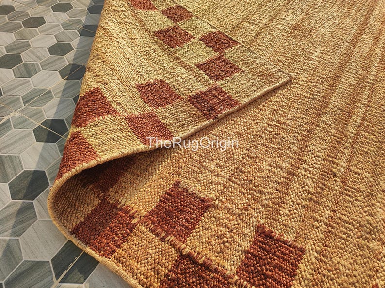 Pu&ograve; includere: Un tappeto in iuta intrecciata con un motivo geometrico nei toni del beige e del marrone rossiccio. Il bordo del tappeto &egrave; ripiegato, rivelando il lato inferiore. Il testo "TheRugOrigin" &egrave; visibile sul tappeto.