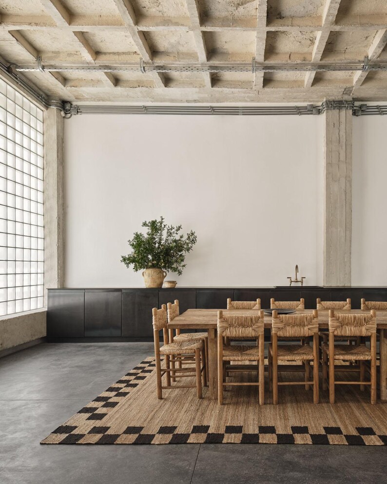 Pu&ograve; includere: Sala da pranzo con tavolo e sedie in legno con sedute intrecciate. Un tappeto rettangolare con un motivo a scacchiera nero e beige &egrave; sotto il tavolo. Una grande pianta in un vaso intrecciato.
