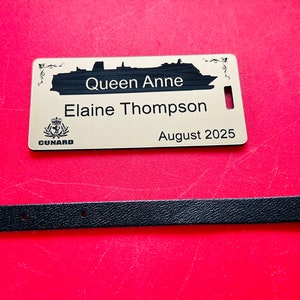 May include: A gold luggage tag with the text "Queen Anne", "Elaine Thompson", and "August 2025". A black leather strap with a silver buckle is also present. The tag features a ship silhouette and the Cunard logo.