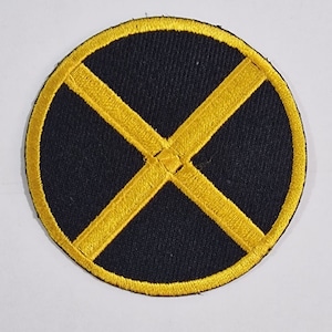 Op de afbeelding: Een ronde, zwarte stoffen patch met een gele X in het midden. De patch is ongeveer 5 cm in diameter.