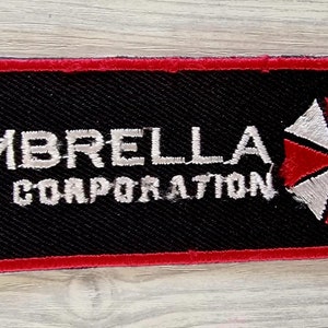 Könnte beinhalten: Ein schwarzer und roter gestickter Aufnäher mit dem Text "Umbrella Corporation" und einem weißen und roten Regenschirm-Logo.