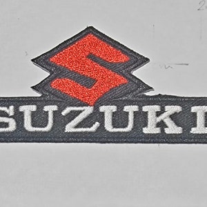 May include: A black and red embroidered patch with the word "SUZUKI" in white lettering. The Suzuki logo is a red "S" in a black diamond shape.
