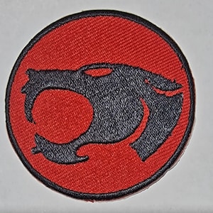 Może przedstawiać: Okrągła, haftowana naszywka w kolorze czerwonym z haftowanym czarnym siluetem głowy lwa.