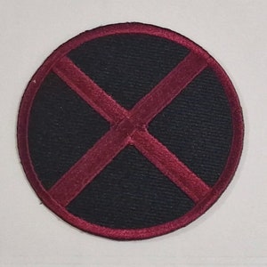 Op de afbeelding: Een ronde, zwarte stoffen patch met een rood geborduurd X in het midden. De patch heeft een rode rand.