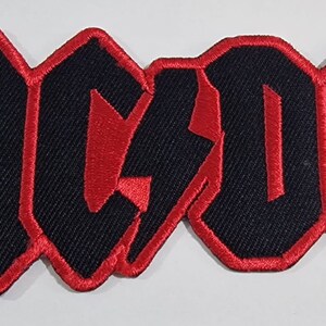 May include: Black and red embroidered patch with the word "AC/DC" in a bold, stylized font. The letters are outlined in red and the lightning bolt is red.