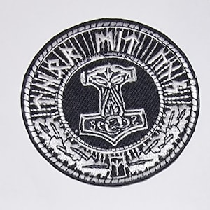 May include: Black and white embroidered patch with a circular design. The center features a stylized hammer with a circle and a design resembling a knot. The outer circle has a repeating pattern of runes and a border.