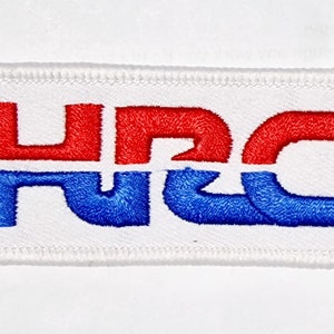 Puede incluir: Un parche blanco bordado con las letras "NRC" en rojo y azul. Las letras están entrelazadas y forman un diseño único.