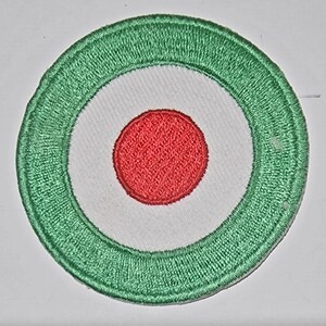 Può includere: Una toppa rotonda ricamata in verde, bianco e rosso. La toppa ha un centro bianco con un cerchio rosso al centro. Il centro bianco è circondato da un anello verde.