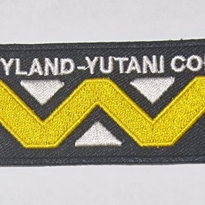Puede incluir: Parche rectangular negro con el texto "WEYLAND-YUTANI CORP" en blanco. Un diseño de chevron amarillo y blanco está centrado debajo. El parche es probablemente un emblema bordado.