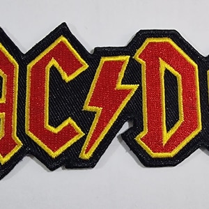 Könnte beinhalten: Ein gestickter Aufnäher in Schwarz, Rot und Gelb mit dem Bandnamen "AC/DC" in einer fetten, stilisierten Schrift.