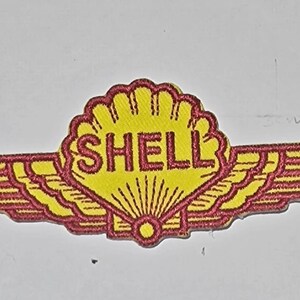 May include: A yellow and red embroidered patch with a stylized shell and the word "SHELL" in red lettering. The shell has a red outline and is surrounded by red wings.