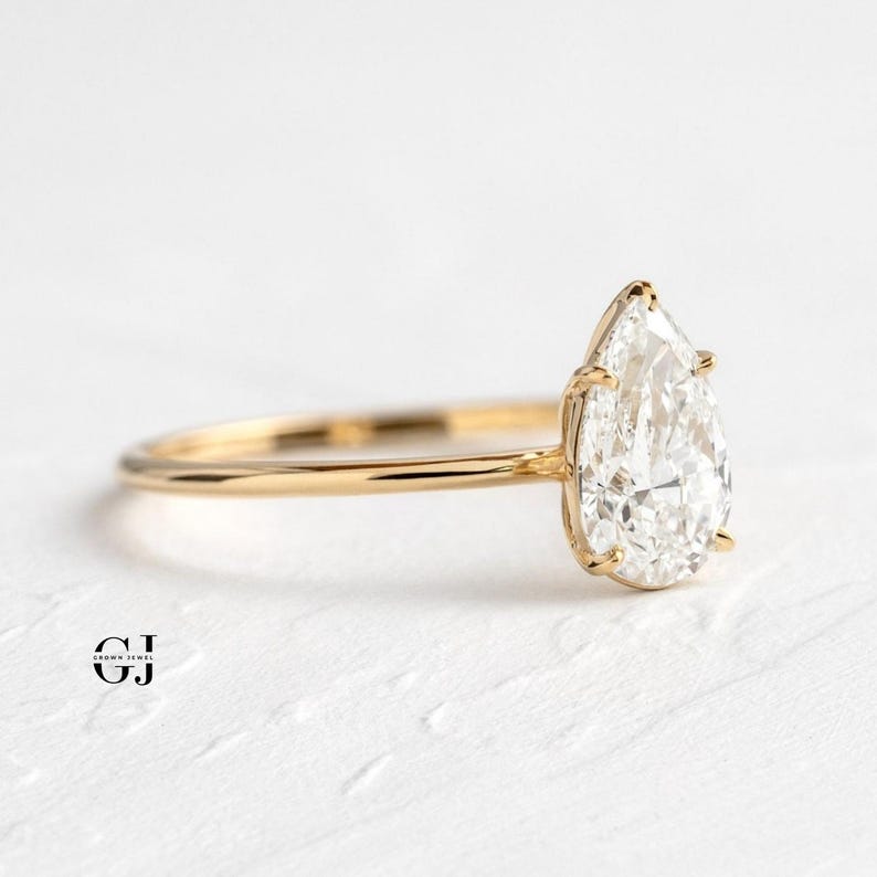 Puede incluir: Un anillo de compromiso de oro con un diamante de talla pera. El diamante es claro y brillante.
