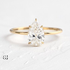 Puede incluir: Un anillo de compromiso de diamante de forma pera engastado en una banda de oro amarillo. El anillo est&aacute; sobre una superficie blanca.