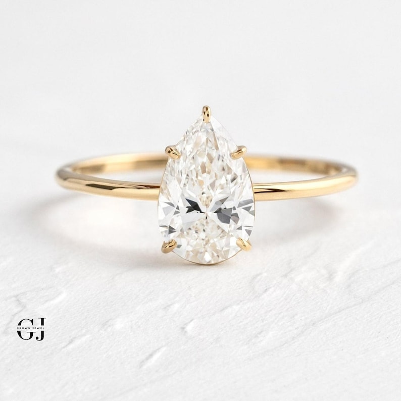 Puede incluir: Un anillo de compromiso de diamante de forma pera engastado en una banda de oro amarillo. El anillo est&aacute; sobre una superficie blanca.