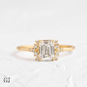 Könnte beinhalten: Ein goldener Verlobungsring mit einem großen, quadratischen, smaragdgeschliffenen Diamanten. Der Diamant ist von einem Halo aus kleineren Diamanten umgeben.