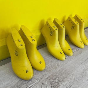 Puede incluir: Seis hormas de zapatos de color amarillo brillante, numeradas con el número 37, están dispuestas contra una pared amarilla. Las formas de madera, utilizadas para dar forma a los zapatos, están sobre un suelo de madera gris claro. La escena evoca un taller.