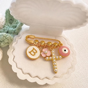 May include: A gold-tone safety pin brooch featuring a white circular charm with the letter B, a small pink flower charm, a cross charm with white beads, and a pink evil eye charm, all resting inside a white plastic shell-shaped container.