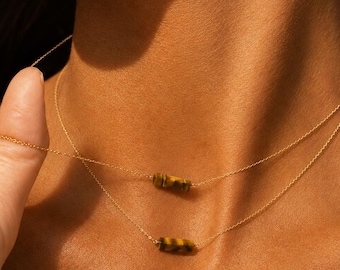 May include: Two gold necklaces with small, cylindrical tiger's eye pendants. The necklaces are layered, with one slightly lower than the other. The chains are delicate, and the pendants have a warm, brown and gold striped pattern.