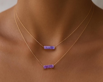 May include: Two gold necklaces with rectangular, lavender-colored pendants. The necklaces are layered, with one pendant slightly above the other. The chains are delicate, and the pendants have a textured appearance.