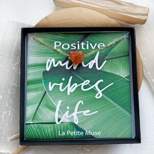 K&ouml;nnte beinhalten: Eine kleine schwarze Box mit einer gr&uuml;nen und wei&szlig;en Karte darin. Die Karte zeigt den Text "Positive mind vibes life" und "La Petite Muse". Eine goldene Kette mit einem orangefarbenen Karneol-Stein liegt auf der Karte.