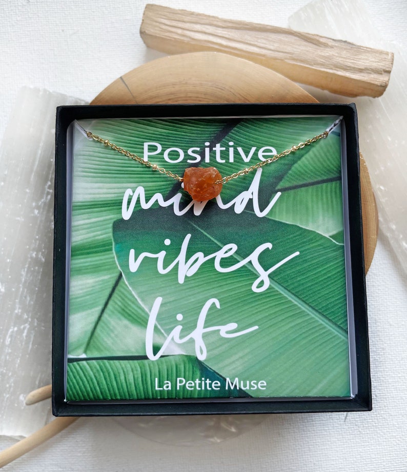 K&ouml;nnte beinhalten: Eine kleine schwarze Box mit einer gr&uuml;nen und wei&szlig;en Karte darin. Die Karte zeigt den Text "Positive mind vibes life" und "La Petite Muse". Eine goldene Kette mit einem orangefarbenen Karneol-Stein liegt auf der Karte.