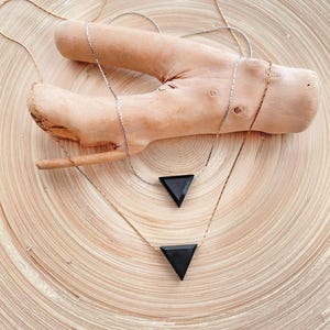 May include: Two necklaces with black triangle pendants. One necklace has a gold chain, the other a silver chain. The necklaces are displayed on a piece of driftwood, set against a light brown, circular background.
