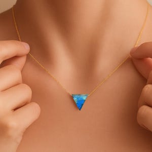 May include: A gold-colored necklace with a triangular pendant featuring shades of blue. The pendant is suspended from a delicate chain. The necklace is being held by a person.