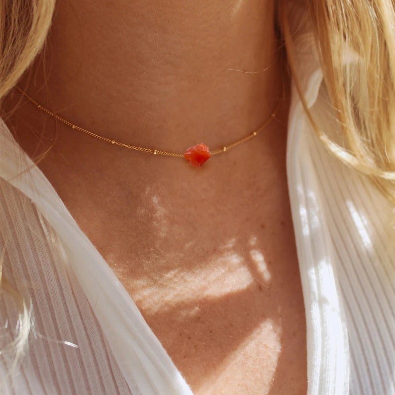 K&ouml;nnte beinhalten: Eine zarte goldene Kettenkette mit einem kleinen, rot-orangenen Edelstein-Anh&auml;nger.