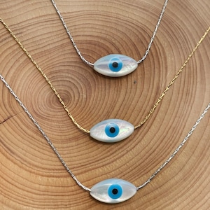May include: Three silver chains with a white oval pendant featuring a blue evil eye charm. The chains are layered on a wooden surface.
