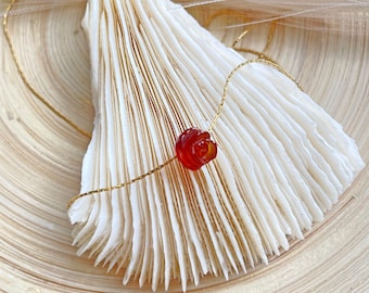 May include: A delicate gold chain necklace with a red rose-shaped pendant. The pendant is made of carnelian, a semi-precious gemstone known for its warm, reddish-orange hue.