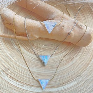 May include: Three delicate necklaces with triangular pendants. The pendants have a shimmering, iridescent quality, with colors of blue, white, and hints of purple. The chains are gold and silver, and the necklaces are displayed on a piece of driftwood.
