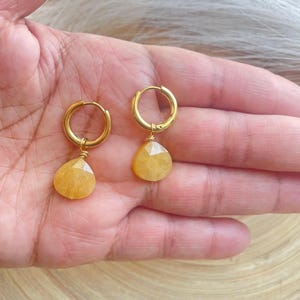 May include: A pair of gold hoop earrings with teardrop-shaped yellow gemstones. The earrings are displayed on a hand, showcasing the polished gold and the vibrant color of the gemstones. The earrings are a jewelry item.