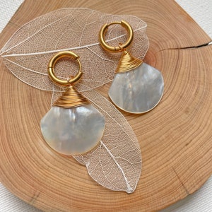May include: A pair of gold hoop earrings with a white shell pendant. The earrings are on a wooden surface with a white leaf.