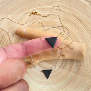 May include: Two delicate necklaces with black triangle pendants. One necklace has a gold chain, the other a silver chain. The pendants are held by a person's finger, set against a wooden background.
