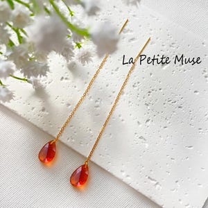 May include: A pair of gold-tone threader earrings featuring delicate chains and faceted, teardrop-shaped orange-red crystals. The text La Petite Muse is visible on the white textured surface near the jewelry pieces.