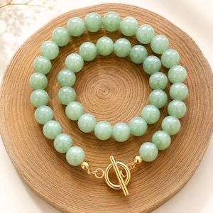 May include: A necklace composed of two strands of smooth, round, pale green jade beads. The strands are connected by a polished gold-tone metal toggle clasp, featuring a circular ring and a bar, with small gold-tone spacer beads at the ends.