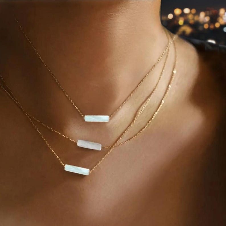 May include: Three gold necklaces with a white bar pendant on each. The necklaces are layered on a person's neck.