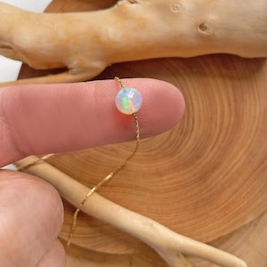 May include: A delicate gold chain necklace with a single round opal pendant. The opal is iridescent with shades of white, blue, and green.