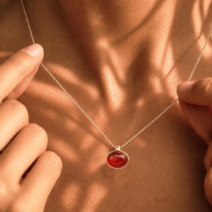 May include: A gold-colored necklace with a red oval pendant. The pendant is set in a gold bezel and hangs from a delicate chain. The necklace is displayed on a person's chest.