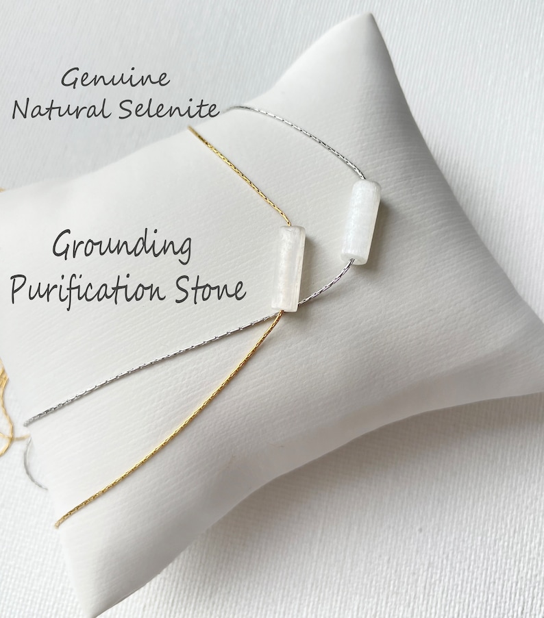 May include: A white selenite grounding and purification stone necklace with a silver and gold chain. The necklace is displayed on a white cushion with the text "Genuine Natural Selenite" and "Grounding Purification Stone".