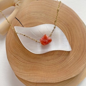 K&ouml;nnte beinhalten: Eine goldene Kettenkette mit einem rohen Karneol-Steinanh&auml;nger. Der Stein ist tief orangefarben und hat eine raue, nat&uuml;rliche Textur. Die Kette wird auf einer Holzoberfl&auml;che pr&auml;sentiert.