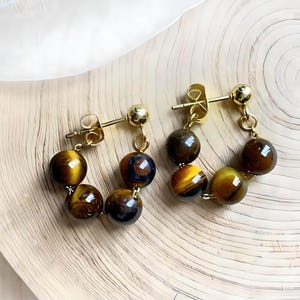 May include: Gold hoop earrings with tiger's eye beads. The earrings are made of gold-toned metal and feature a chain of tiger's eye beads.