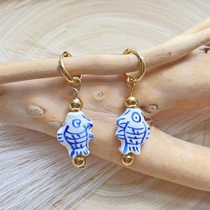 May include: A pair of gold hoop earrings with blue and white porcelain fish charms. The fish charms have a blue and white design.