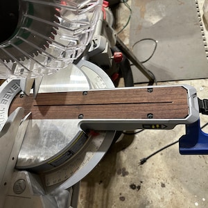 May include: A close-up of a miter saw with a wooden extension attached to the base. The extension has two grooves and is secured with screws. The saw has a clear plastic guard and a blue lever.